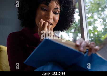 Rücksichtsvolle, birassische Frau, die am Fenster sitzt und zu Hause ein Buch liest Stockfoto