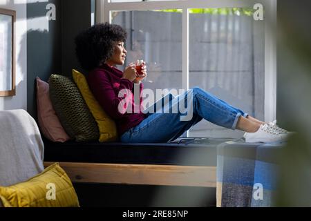 Rücksichtsvolle, birassische Frau, die am Fenster sitzt und zu Hause Tee trinkt Stockfoto
