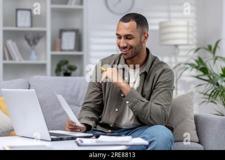Junger afroamerikanischer Mann, der zu Hause mit Laptop und Dokumenten arbeitet. Hält und liest einen Brief, eine Vereinbarung, eine Rechnung, lächelt zufrieden. Stockfoto