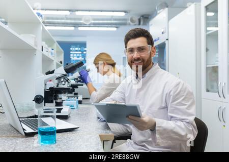 Porträt eines jungen männlichen Wissenschaftlers, der mit einem Lächeln in die Kamera schaut. Er sitzt im Labor vor einem Mikroskop mit Dokumenten, eine Kollegin im Hintergrund. Stockfoto