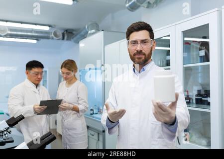 Porträt eines lächelnden jungen Mannes, der einen Plastikbehälter mit einer Substanz in die Kamera hält. Er arbeitet in einem Labor für die Herstellung von Kosmetika und Haushaltschemikalien. Stockfoto