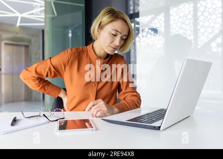 Müde und überarbeitete Frau, die im Büro arbeitet, Geschäftsfrau hat starke Rückenschmerzen, verärgerte blonde Muskelmasseur, lange sitzende Arbeit. Stockfoto