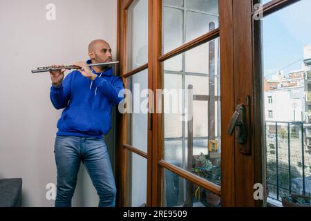 Junger Latino-Mann in blau gekleidet, allein zu Hause, spielt Flöte, schaut aus dem Fenster und denkt mit einem melancholischen Gesicht. People Concept, kopiere Spa Stockfoto