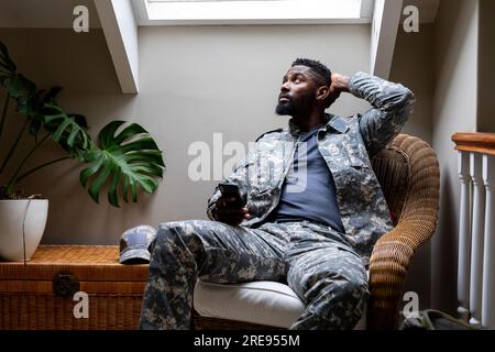 afroamerikanischer Soldat in Militäruniform, der zu Hause sitzt und ein Smartphone benutzt Stockfoto