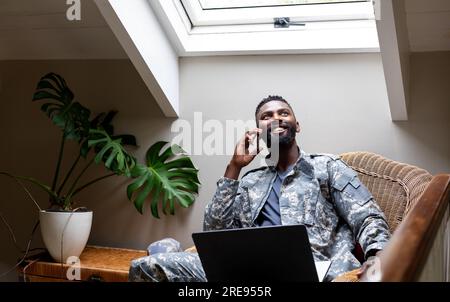 afroamerikanischer Soldat trägt Militäruniform mit Laptop und spricht zu Hause über Smartphone Stockfoto