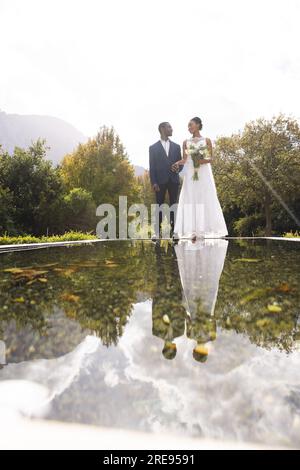 Fröhlicher afroamerikanischer Bräutigam und Braut, die einen Blumenstrauß im Teich im sonnigen Garten halten, Kopierbereich Stockfoto