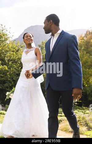 Glückliche afroamerikanische Braut und Bräutigam, die Händchen hielten und im sonnigen Garten spazieren gingen Stockfoto