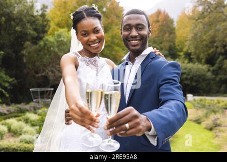 Porträt einer glücklichen afroamerikanischen Braut und Bräutigam, die mit Champagner im sonnigen Garten anstoßen Stockfoto