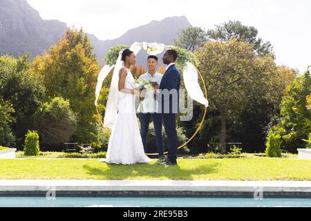 Ein vielseitiger männlicher Beamter, Braut und Bräutigam bei der Hochzeitszeremonie im Freien am Pool im sonnigen Garten Stockfoto