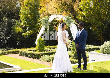 Glückliche afroamerikanische Braut und Bräutigam stehen unter dem Hochzeitsbogen im sonnigen Garten, Kopierbereich Stockfoto