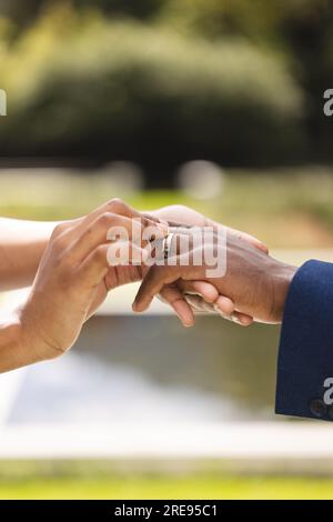 Hände einer afroamerikanischen Braut, die den Ring auf den Finger des Bräutigams im sonnigen Garten legt, Kopierraum Stockfoto