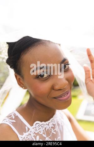 Porträt einer lächelnden afroamerikanischen Braut mit weißem Schleier bei der Hochzeitszeremonie im sonnigen Garten Stockfoto