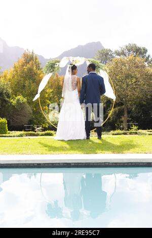 Rückblick auf afroamerikanische Braut und Bräutigam unter dem Hochzeitsbogen am Pool im sonnigen Garten, Kopierbereich Stockfoto