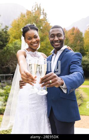 Porträt einer glücklichen afroamerikanischen Braut und Bräutigam, die mit Champagner im sonnigen Garten anstoßen Stockfoto