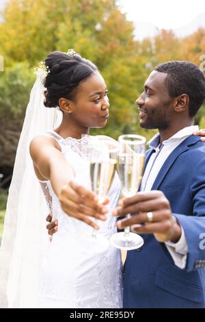 Frohe afroamerikanische Braut und Bräutigam, die sich im sonnigen Garten mit Champagner anlächelt Stockfoto