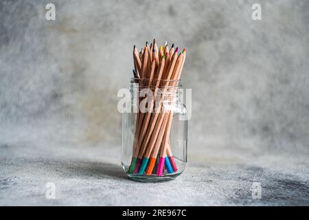 Sammlung von bunten Bleistiften, die in einem Glasbecher gestapelt und vor grauem Hintergrund auf dem Tisch platziert sind Stockfoto