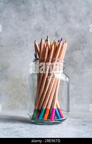 Sammlung von bunten Bleistiften, die in einem Glasbecher gestapelt und vor grauem Hintergrund auf dem Tisch platziert sind Stockfoto