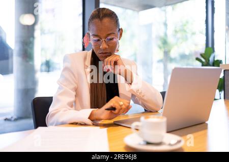 Nachdenkliche afroamerikanische Geschäftsfrau mit langen Haaren in einem formellen weißen Blazer, die mit Kaffee und Laptop am Tisch sitzt Stockfoto