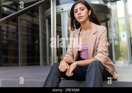 Kleiner Winkel einer selbstbewussten Unternehmerin in formellen Outfits, die auf Treppen in der Nähe des Eingangs des Bürogebäudes sitzt und auf die Kamera schaut Stockfoto