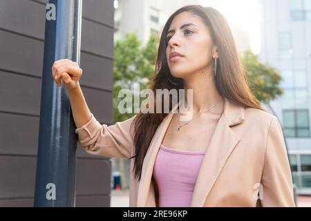 Nachdenkliche junge hispanische Unternehmerin in formeller Kleidung, die auf der Straße neben der Metallsäule steht und wegschaut Stockfoto