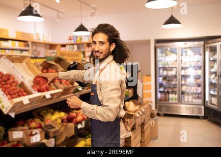 Seitenansicht eines glücklichen, ethnischen, männlichen Verkäufers mit lockigem Haar in Uniform, der während der Arbeit im Supermarkt frische reife Tomaten aus einer Holzkiste pflückt Stockfoto
