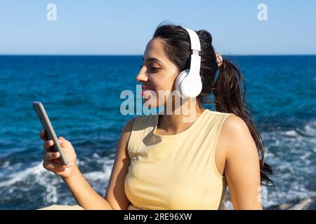 Lächelnde indische Frau in legerer Kleidung mit kabellosen Kopfhörern, die am sonnigen Tag am Meer auf dem Handy surft Stockfoto