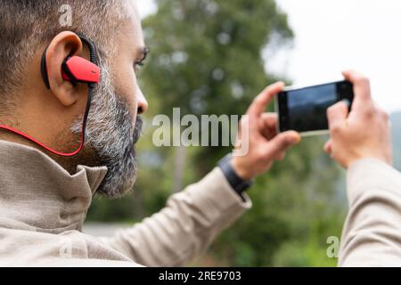 Seitenansicht eines bärtigen indischen Mannes, der auf den Bildschirm des Mobiltelefons schaut und ein Foto macht, während er an sonnigen Tagen mit TWS-Ohrstöpseln am Nackenbügel steht Stockfoto