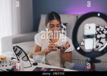 Junge hispanische Influencer, die Bronzer mit Pinsel aufträgt, während sie Make-up-Tutorial auf Smartphone in einem modernen Raum aufnimmt Stockfoto