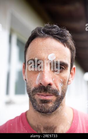 Porträt eines bärtigen männlichen Mannes in legerer Kleidung mit weißen Farbtropfen auf dem Gesicht, der in die Kamera schaut, während er gegen das Äußere eines verschwommenen Gebäudes steht Stockfoto