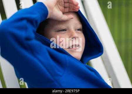 Lächelndes Kind im Hoodie, das wegschaut, während es draußen auf dem Stuhl sitzt und sich im Sommer vor der unscharfen grünen Natur versteckt Stockfoto