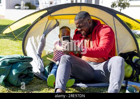 Fröhlicher afroamerikanischer Sohn und Vater, der Selfie macht und im sonnigen Zeltgarten sitzt Stockfoto