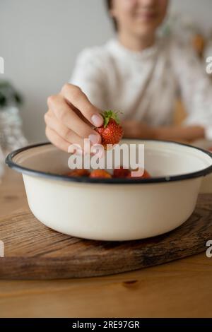 Nahaufnahme einer Schüssel frischer Erdbeeren und einer Hand anonymer junger Frau, die Erdbeeren nimmt, während sie mit verschwommenem Hintergrund am Tisch sitzt Stockfoto