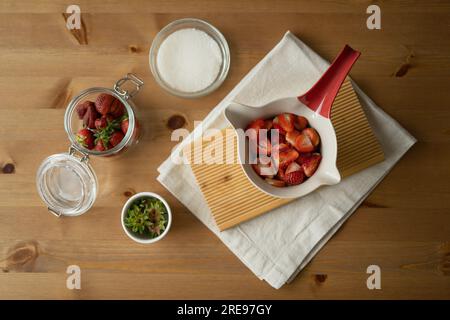 Blick von oben auf köstliche, frisch geschnittene Erdbeeren und Zucker in Glas- und Keramikschüssel auf Holztisch mit Stoff während der Konfitüre im Innenbereich Stockfoto