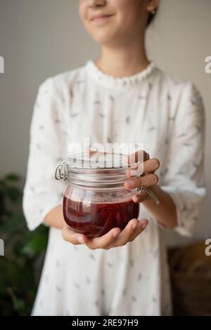 Pflücken Sie eine anonym positive Frau in weißem, legerem Tuch, halten Sie ein Glas Erdbeermarmelade und lächeln Sie, während Sie vor dem verschwommenen Hintergrund des Raumes stehen Stockfoto