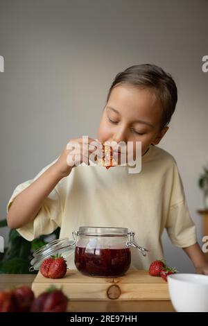 Porträt eines traumhaften Mädchens in legerer Kleidung, das köstlichen Pfannkuchen mit hausgemachter Erdbeermarmelade isst, während er in einem hellen Raum am Tisch steht Stockfoto