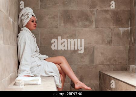 Seitlicher Blick auf die junge Frau im weißen Bademantel auf dem Kopf, während sie während des Urlaubs in einem gefliesten Hamam sitzt Stockfoto
