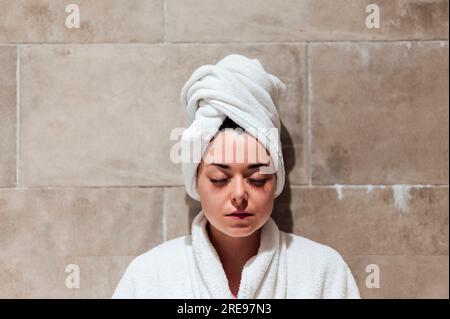 Eine junge Frau im weißen Bademantel am Kopf, die während des Urlaubs mit geschlossenen Augen im gefliesten Hamam sitzt Stockfoto