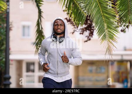 Fokussierter junger afroamerikanischer Mann mit Bart in Sportbekleidung schaut in die Kamera, während er bei Tageslicht auf der Straße in der Nähe eines Gebäudes läuft Stockfoto