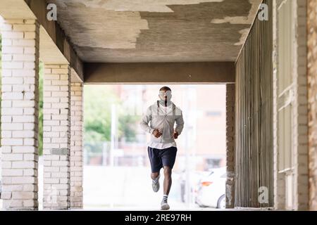 Fokussierter junger afroamerikanischer Mann mit Bart in Sportbekleidung schaut in die Kamera, während er auf dem Boden der Veranda mit Ziegelsäulen und Dach in da joggt Stockfoto