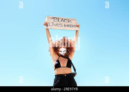 Niedriger Winkel der ethnischen Frau in der medizinischen Maske stehen mit Plakat mit Rückseite lebt Materie Inschrift auf Hintergrund blau Wolkenloser Himmel Stockfoto