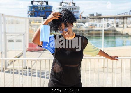 Entzückte Frau in trendigen tragen und mit afro Frisur stehen Am Kai mit Schutzmaske am sonnigen Tag und Blick Weg Stockfoto
