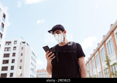 Von unten von unerkennbarem Männchen in Gesichtsmaske und Mütze Surfen im Internet auf Handy unter Himmel während COVID 19 Pandemie Stockfoto