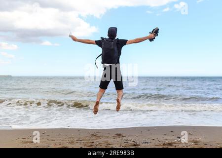 Rückansicht des anonymen Inhalts männlicher Reisender mit Rucksackspringen In der Luft mit erhobenen Armen über sandigen Ufer in der Nähe des Ozeans Unter blauem bewölktem Himmel Stockfoto