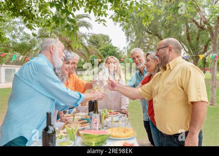 Gruppe positiver männlicher und weiblicher Freunde mittleren Alters, die während des Geburtstags im Park mit Weingläsern klappern Stockfoto