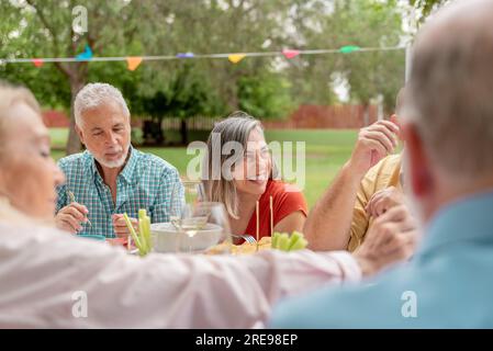 Eine Gruppe von Freunden mittleren Alters, die verschiedene Speisen und Wein zu sich nehmen, während sie ihren Geburtstag im Park feiern Stockfoto