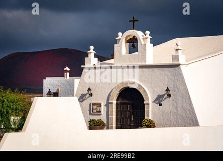 Eine weiß getünchte Kirche auf Lanzarote mit stürmischem Himmel und dem Vulkan Caldera Colorada im Hintergrund Stockfoto