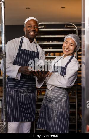 Porträt von fröhlichen, vielfältigen Bäckern, die Schürzen tragen und Tafel in einer Bäckerei mit überkreuzten Armen verwenden Stockfoto