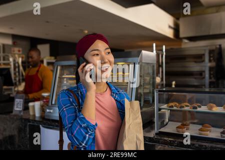 Zufriedener, vielseitiger männlicher Bäckereiangestellter und Kunde mit Papiertüte, die auf dem Smartphone spricht Stockfoto