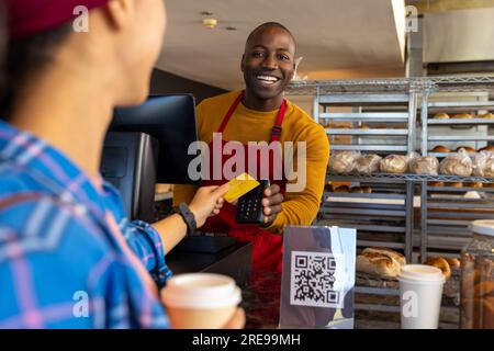 Glücklicher, vielseitiger männlicher Bäckereiangestellter und Gast, der mit Kreditkarte bezahlt Stockfoto