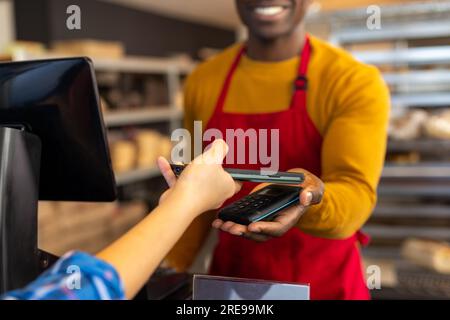 Zufriedener, vielseitiger männlicher Bäckereiarbeiter und Kunde, der mit einem Smartphone mit Kopierbereich bezahlt Stockfoto